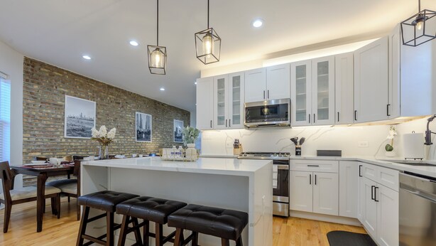 A modern kitchen with white cabinets, a central island with stools, a brick accent wall, and hanging pendant lights. - Queen Beds, EV Charging, Near Train: Luxury Stay (Chicago)