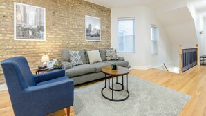 A modern living room with a brick wall, a grey sofa, a blue armchair, and a round wooden coffee table. - Queen Beds, EV Charging, Near Train: Luxury Stay (Chicago)