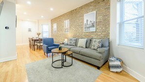 A modern living room with a brick accent wall, a grey sofa, a blue armchair, and a dining area with wooden flooring and a table. - Queen Beds, EV Charging, Near Train: Luxury Stay (Chicago)