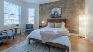 A modern bedroom with a brick wall, a large bed, a chair, and a nightstand. - Queen Beds, EV Charging, Near Train: Luxury Stay (Chicago)