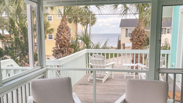 A covered outdoor area with two white chairs and a small table, overlooking a beach with palm trees and buildings in the background. - Gentleman Pirate (Ocean Views!) (Edisto Island)