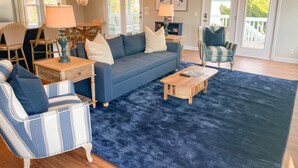 A living room with a blue sofa, two armchairs, a coffee table, and a rug. There are two lamps, a bookshelf, and a door leading to a balcony. - Gentleman Pirate (Ocean Views!) (Edisto Island)