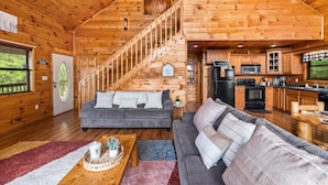 A spacious cabin with a wooden interior, a staircase, a living room with two sofas, a kitchen area, and a dining table with a tray.