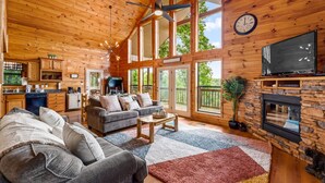 A spacious living room with a large sofa, a coffee table, and a fireplace. There is a kitchen area with a dishwasher and a grill. The room has large windows and a wooden ceiling.