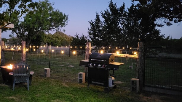 Un patio exterior con una parrilla, sillas y luces colgantes.