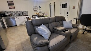 Una sala de estar moderna con un sofá de cuero gris, un comedor y, al fondo, una cocina, además de una silla de oficina negra.