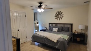 Un dormitorio con cama, ventilador de techo, reloj de pared y mesita de noche con lámpara.