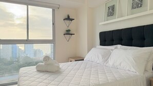 A bedroom with a large window, a bed with white bedding, two wall-mounted shelves with plants, and a city view. - Ocean View 1BR - TV 65" - Pool - Jacuzzi 1606 (Barranco)
