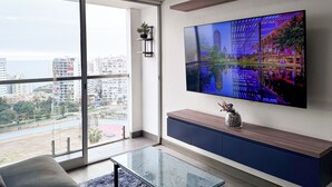 A modern living room with a large TV, a glass coffee table, a sofa, and a view of a cityscape through a large window. - Ocean View 1BR - TV 65" - Pool - Jacuzzi 1606 (Barranco)