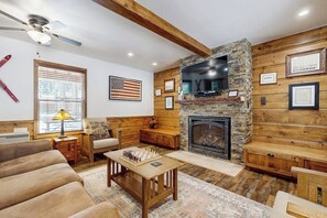 A living room with a fireplace, wooden walls, a ceiling fan, and a chessboard.
