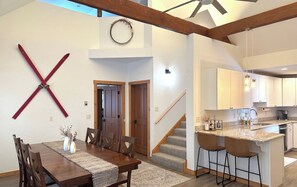 Uma sala de jantar com mesa e cadeiras de madeira, um objeto decorativo na parede e uma escada para outro andar.