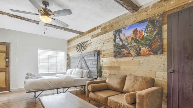 Une pièce avec un plafond en bois, un ventilateur de plafond, un lit avec une literie blanche, un canapé en cuir marron, une table basse en bois et un tableau représentant un paysage de montagne accroché au mur.