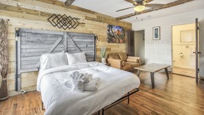 Une chambre à coucher avec une tête de lit en bois, un lit recouvert de draps blancs, un ventilateur de plafond, un tableau accroché au mur et une porte donnant sur une salle de bain.