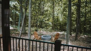 Een houten terras met een metalen reling, twee Adirondack-stoelen en een vuurkorf in een bebost gebied.