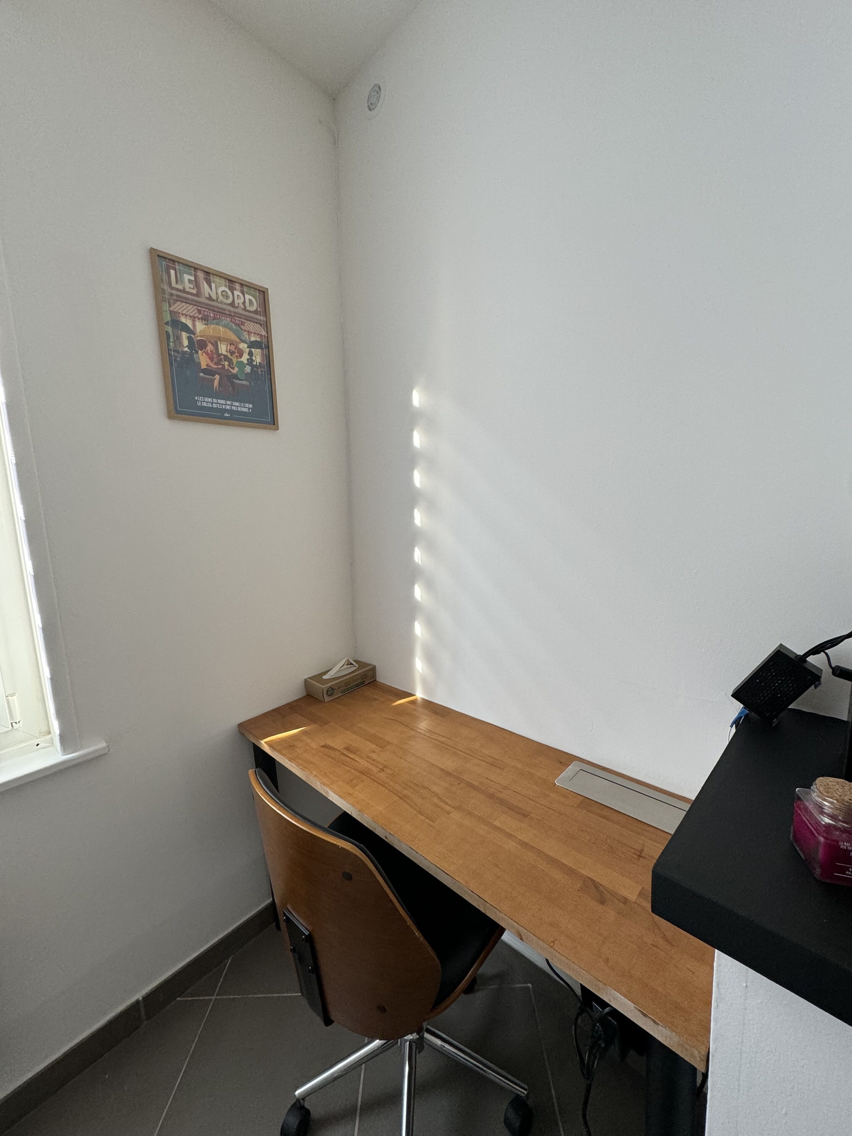 Ein kleiner Raum mit einem Holztisch, einem Stuhl, einem Fenster und einem Poster an der Wand.