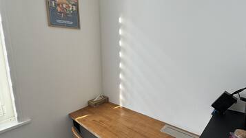 Ein kleiner Raum mit einem Holztisch, einem Stuhl, einem Fenster und einem Poster an der Wand.