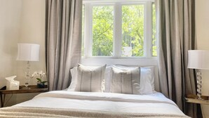 Un lit bien fait, avec du linge de lit blanc et un plaid beige, flanqué de deux tables de chevet sur lesquelles reposent des lampes et une boîte de mouchoirs.