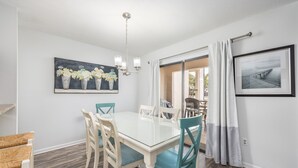 A dining area with a glass table and teal chairs, a chandelier, and a painting of vases on the wall.