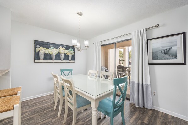A dining area with a glass table and teal chairs, a chandelier, and a painting of vases on the wall.
