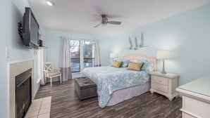 A bedroom with a bed, bedside tables, a TV mounted on the wall, a ceiling fan, and a fireplace.