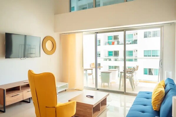 A modern living room with a flat-screen TV, a yellow chair, a blue sofa, and a large sliding glass door leading to an outdoor balcony.