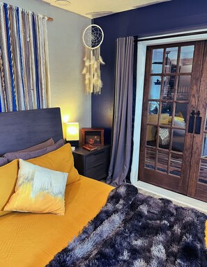 A bedroom with a bed, a nightstand, a dreamcatcher, curtains, and a view through a glass door.