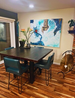 A dining area with a wooden table, four chairs, a large abstract painting, and a potted plant.