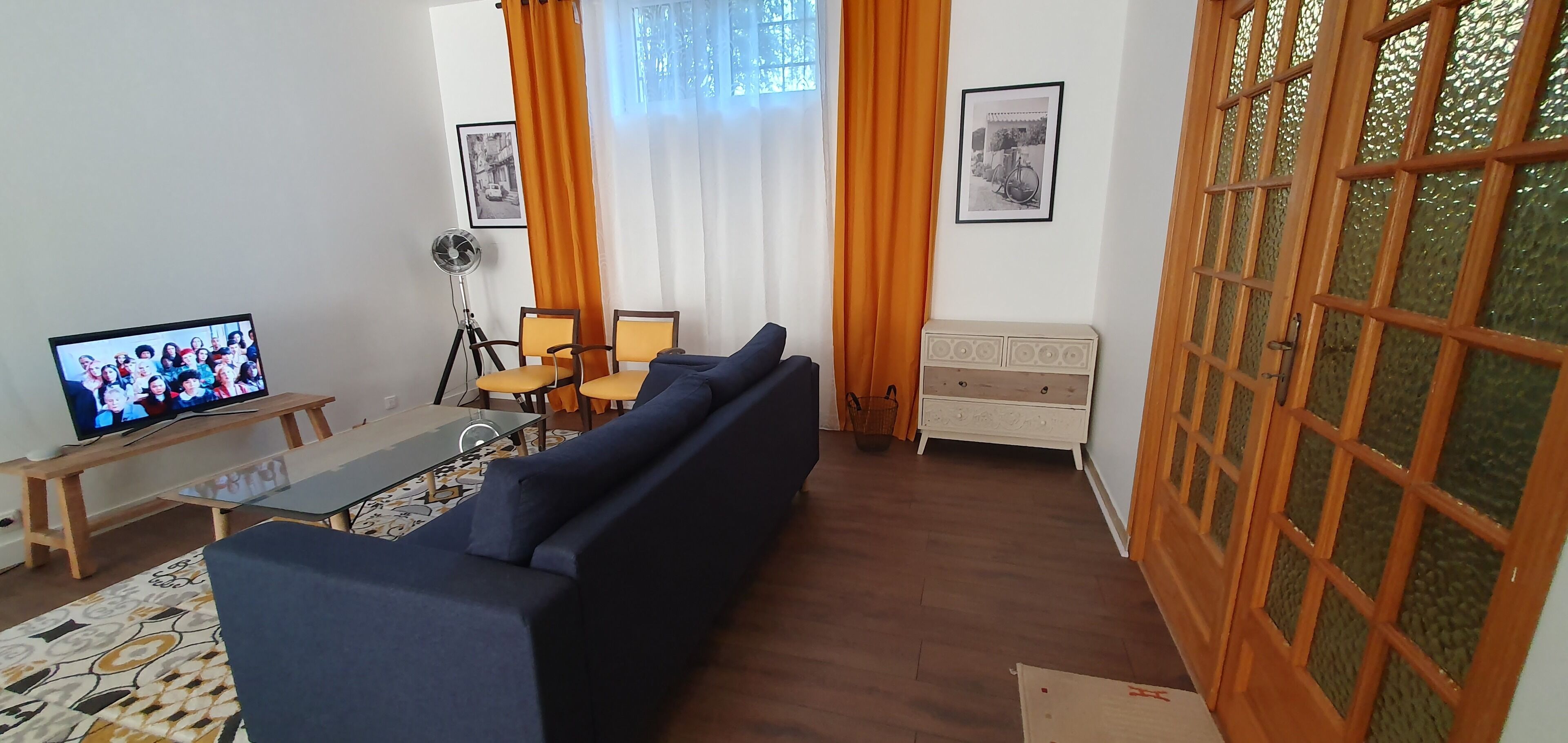 Ein Wohnzimmer mit einem dunkelblauen Sofa, einem Glastisch und einem Fernseher auf einem Holzbodenständer. Zwei gelbe Sessel, ein weißes Sideboard und ein Fenster mit orangenen Vorhängen ergänzen den Raum.