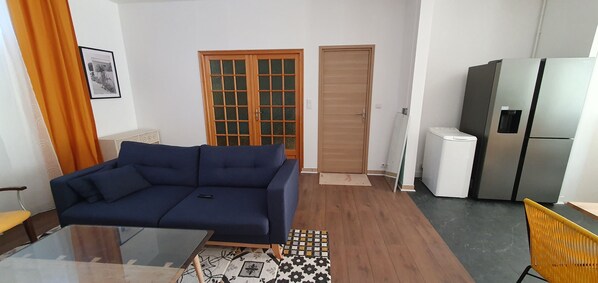 Una sala de estar con un sofá azul, una mesa de centro de vidrio, un tapete estampado, una puerta de madera, un refrigerador y una silla amarilla.