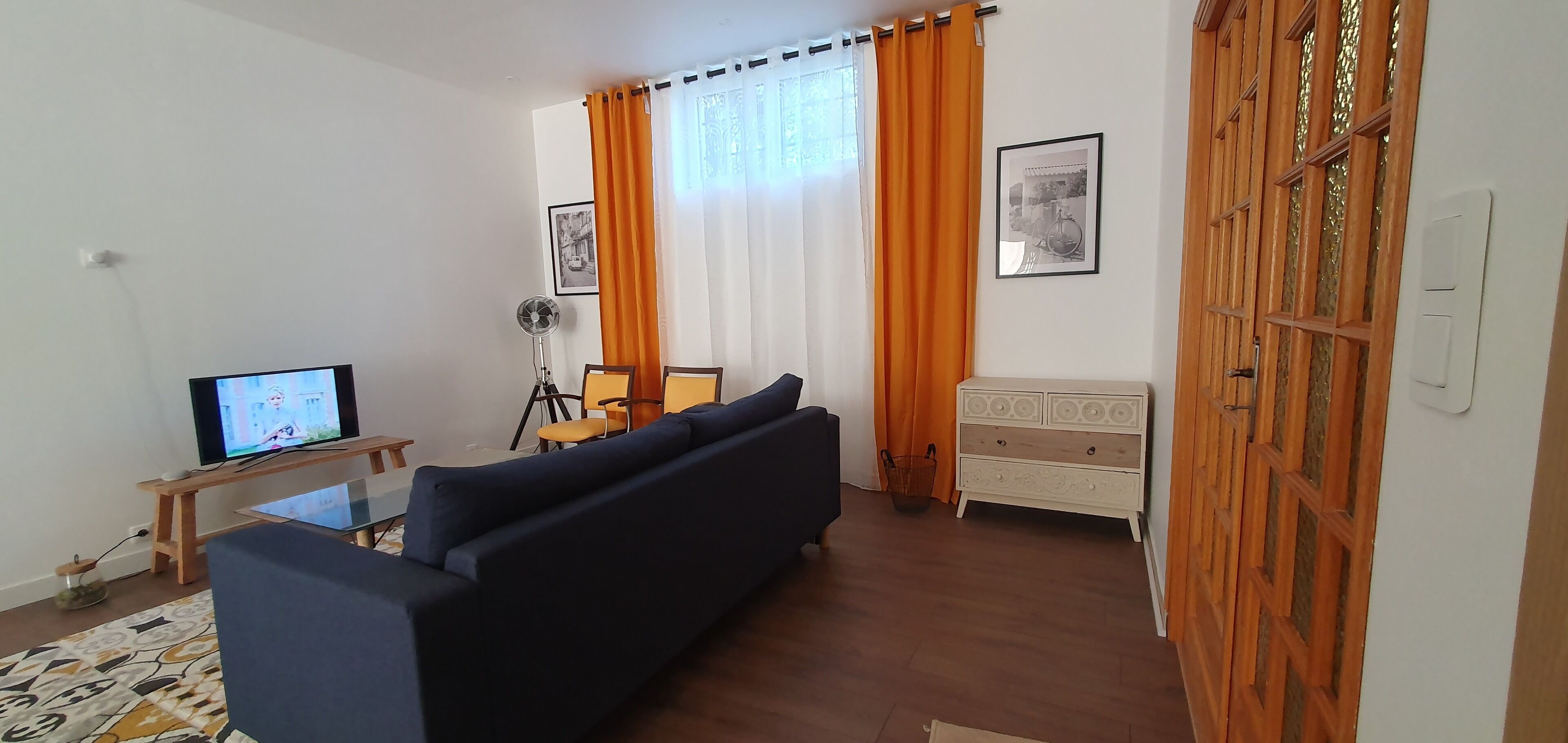 Ein Wohnzimmer mit einem dunklen Sofa, einem Fernseher auf einem Holzständer, einer Stehlampe und einer weißen Kommode mit Spiegel.