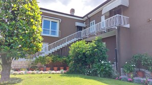 Ein zweistöckiges Gebäude mit weißer Treppe, einem Balkon und einem Garten mit verschiedenen Pflanzen und Blumen.
