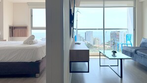 A modern hotel room with a bed, a desk, a sofa, and a large window offering a city view. - Ocean View 1BR - TV 65" - Pool - Jacuzzi 1609 (Barranco)
