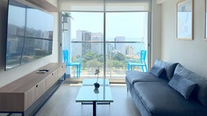 A modern living room with a large flat-screen TV, a grey sofa, a glass coffee table, and a balcony with a view of the city and buildings. - Ocean View 1BR - TV 65" - Pool - Jacuzzi 1609 (Barranco)