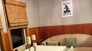 A dining area with a table, chairs, and a candle centerpiece. A sofa is in the background, and a wall sign reads "DO YOU BELIEVE".