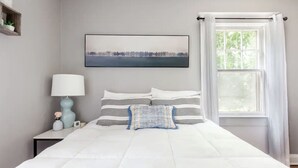 Un dormitorio con una cama grande, una mesita de noche con lámpara, un cuadro en la pared y una ventana con cortinas.