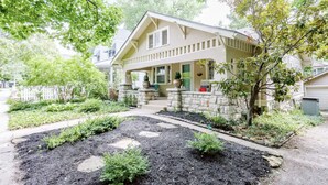 Una casa de dos pisos con cimientos de piedra, un porche delantero y un jardín bien cuidado con acolchado y plantas.