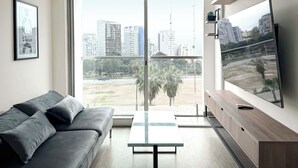 Ein modernes Wohnzimmer mit großem Fenster, einem grauen Sofa, einem Glastisch und einem Flachbildfernseher.