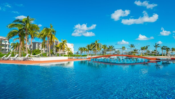 A swimming pool with lounge chairs and palm trees, surrounded by modern buildings. - Sirenita G. Infinity pool - At Mareazul (Playa del Carmen)