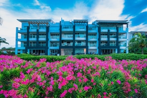 A modern multi-story hotel with balconies and a landscaped garden with pink flowers.
