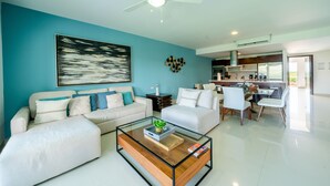 A modern living room with a glass coffee table, white sofas, and a kitchen in the background. - Sirenita G. Infinity pool - At Mareazul (Playa del Carmen)