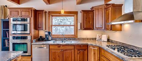 A kitchen with wooden cabinets, a stainless steel range, a dishwasher, and a window with a view of trees.