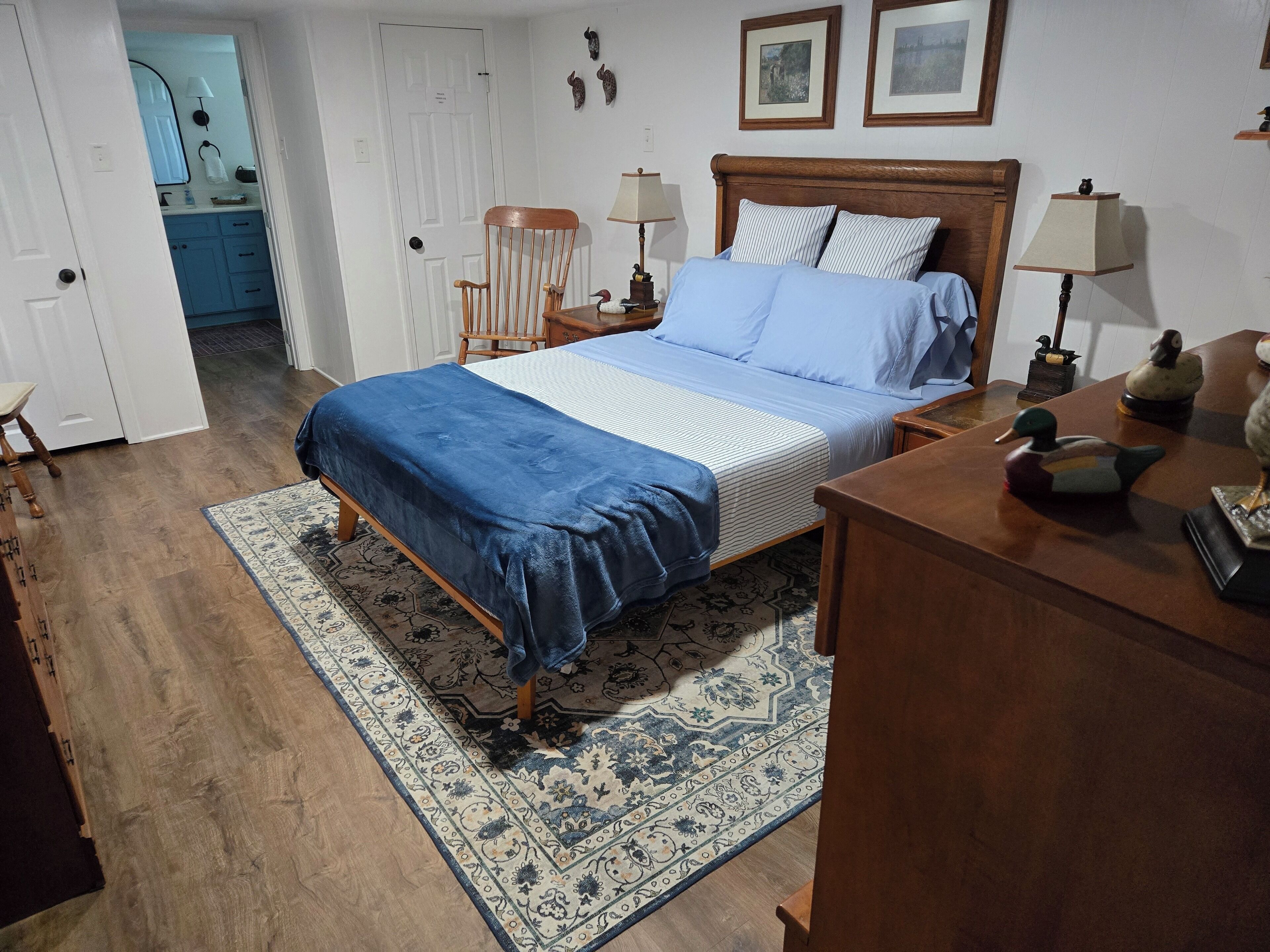 A bedroom with a bed, two bedside tables, a wooden dresser, a patterned rug, and a door leading to a bathroom.