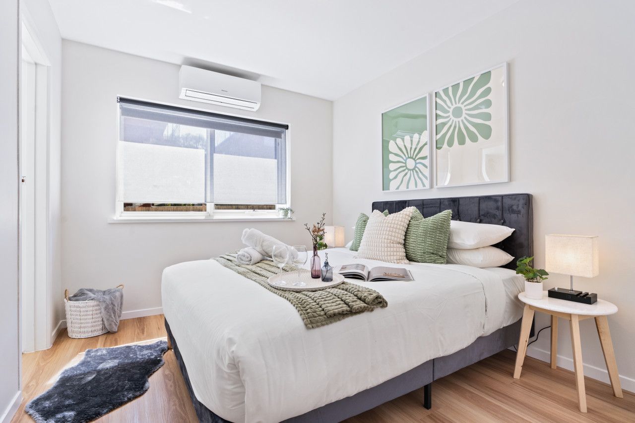 A modern bedroom with a large bed, a bedside table with a lamp, a window with blinds, and a wall-mounted air conditioner.