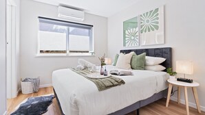 Une chambre moderne avec un grand lit, une table de chevet avec une lampe, une fenêtre avec des stores et un climatiseur fixé au mur.