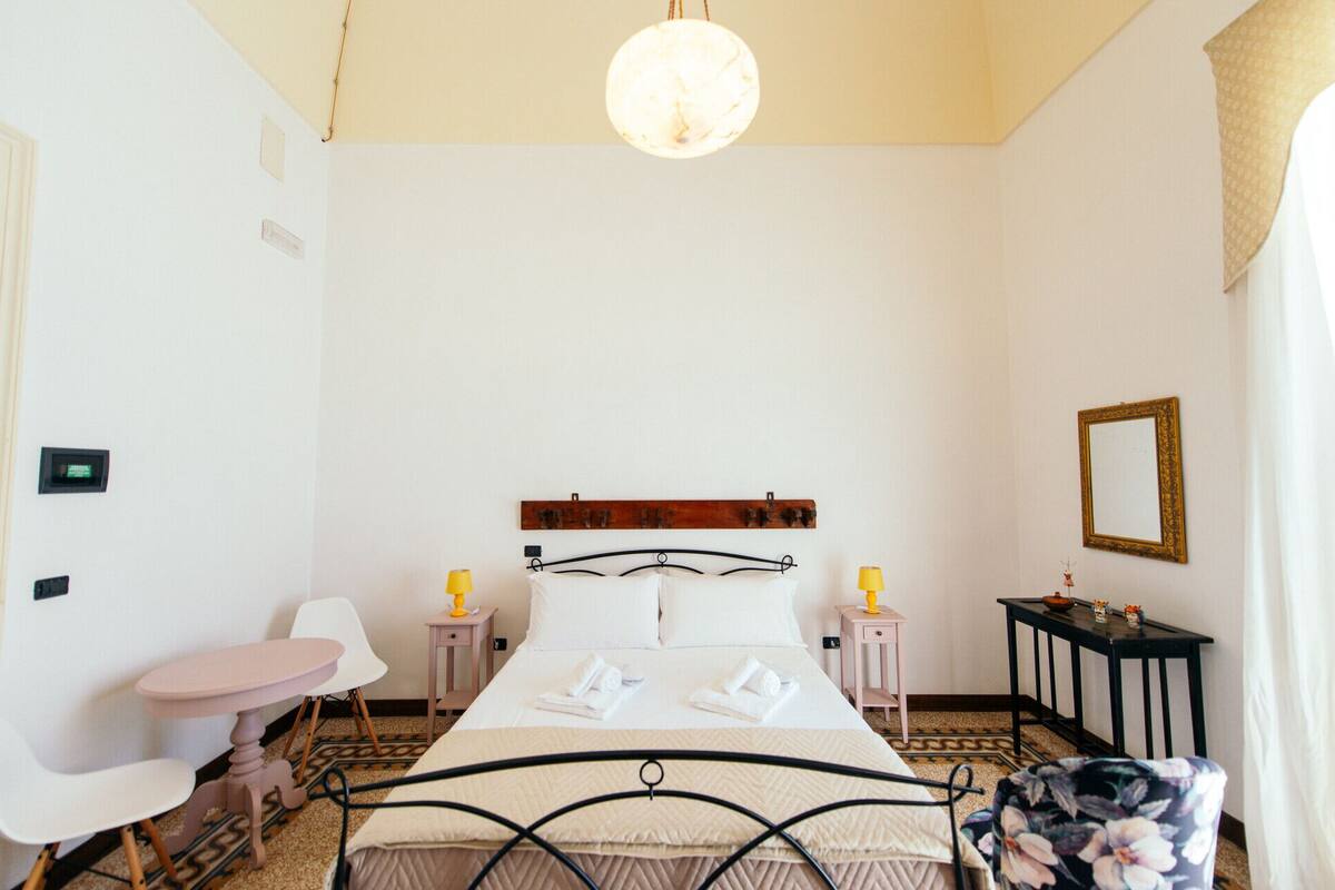 Habitación de hotel con cama, mesita de noche, una silla, una pequeña mesa, espejo y lámpara colgante.