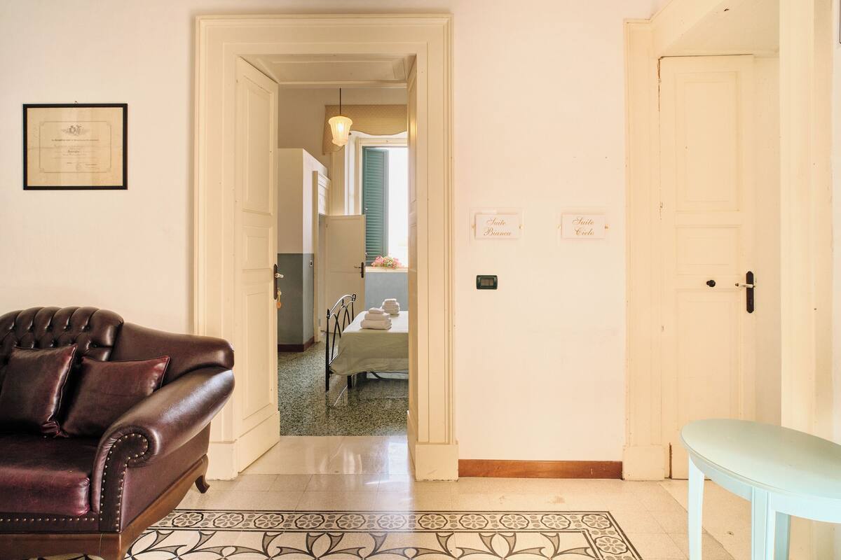 Una habitación con un sofá de cuero marrón, una mesa azul claro y un piso estampado. Hay una puerta que da a otra habitación con una cama y una ventana.