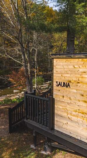 Estructura de sauna de madera con un letrero que dice "SAUNA", ubicada en un área boscosa con un pasillo de madera que la conduce.