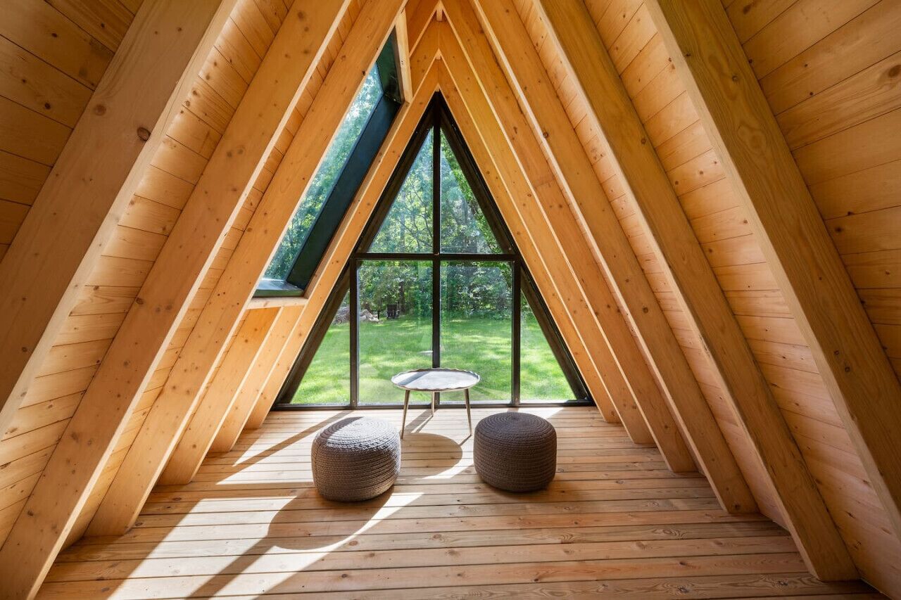 A triangular attic room with wooden beams, a large window, and two round ottomans.