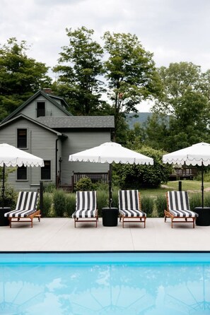 A poolside area with lounge chairs and umbrellas, a house in the background, and a clear blue pool.