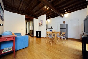 A living room with a blue sofa, wooden dining table, and chairs, a kitchen area with a refrigerator, and a TV on a stand.